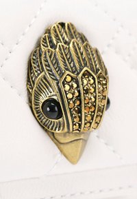 Decorazione metallica a forma di uccello con strass dorati e marroni, piume testurizzate e dettagli degli occhi neri, su uno sfondo bianco trapuntato.