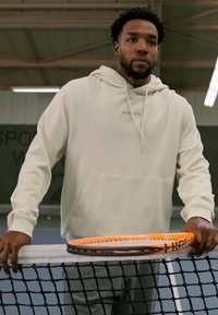 Hooded sweatshirt in soft cream, with a front pocket and subtle branding; grey pants. Holding an orange-accented tennis racket at a net.