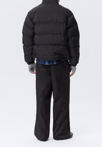 Chaqueta acolchada negra con un cuello alto y secciones acolchadas horizontales, combinada con pantalones negros holgados y guantes sin dedos grises, vista desde atrás.