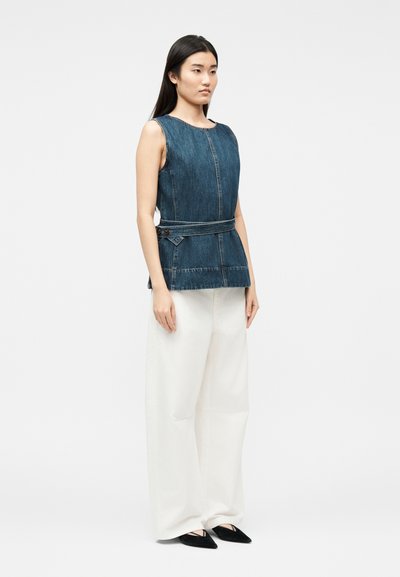 Femme debout portant un haut en denim sombre sans manches avec ceinture, un pantalon blanc à jambes larges et des ballerines noires pointues, légèrement tournée vers la droite.