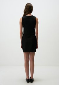 Robe noire sans manches avec une silhouette ajustée, présentant un encolure épurée et une texture lisse, associée à des chaussures noires.