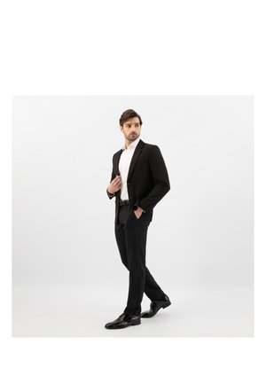 Homme en costume noir, chemise blanche et chaussures habillées noires, debout avec une main dans la poche, regardant vers la gauche sur un fond blanc uni.