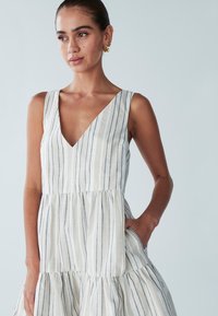 Sukienka bez rękawów w jasno-beżowym kolorze z pionowymi niebieskimi paskami, z dekoltem w kształcie litery V, rozkloszowaną, warstwową spódnicą oraz bocznymi kieszeniami.
