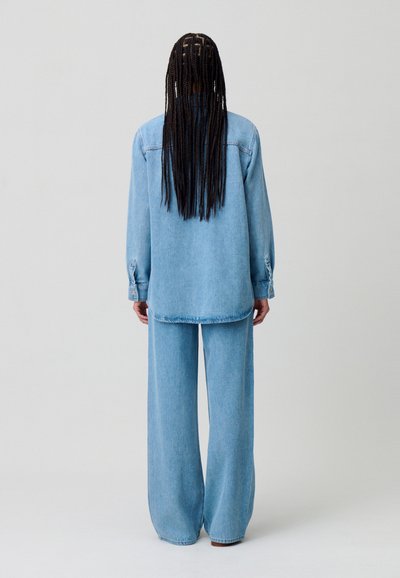 Completo in denim azzurro chiaro composto da una camicia a bottoni dalle linee morbide e pantaloni a gamba larga, con una superficie liscia e dettagli hardware minimi.