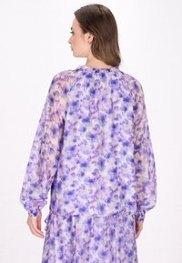 Blusa floreale in tessuto leggero e trasparente con motivi viola e lavanda. Maniche a sbuffo con polsini elastici e scollatura arricciata.