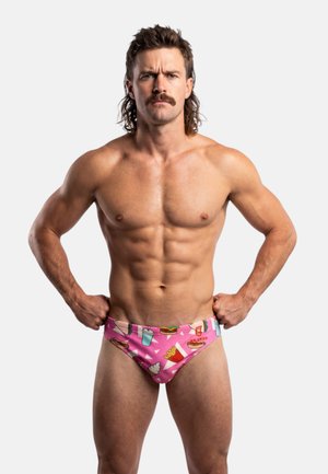 Hombre musculoso con bigote y peinado mullet, de pie con las manos en las caderas, vistiendo un bañador rosa con estampado de comida rápida sobre fondo blanco.