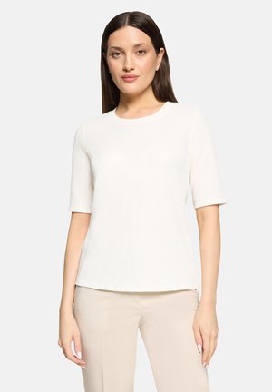 Femme aux cheveux noirs et lisses portant un haut blanc à manches courtes texturé et un pantalon beige, debout devant un fond clair uni.