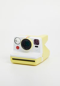 Polaroid camera with yellow plastic body, white front panel, circular lens, red button, and rainbow stripe along the top. Instant photo slot at bottom.