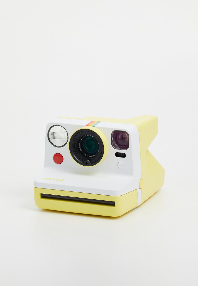 Polaroid camera with yellow plastic body, white front panel, circular lens, red button, and rainbow stripe along the top. Instant photo slot at bottom.