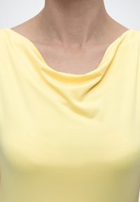Γυναίκα που φοράει ένα ανοιχτό κίτρινο τοπ με λαιμόκοψη κασκόλ που πέφτει, αποκαλύπτοντας την κλείδα και την περιοχή του άνω στήθους.