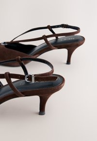 Sandales en suède marron à petit talon avec fines lanières croisées et boucle argentée sur fond clair.