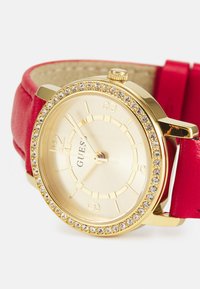 Goldfarbener Uhr mit rundem Ziffernblatt, rotem Lederarmband und Kristallakzenten um die Lünette. Verfügt über ein beiges Zifferblatt mit goldenen Stundenmarkierungen.
