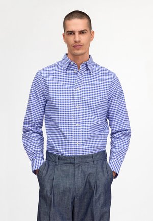 Homme portant une chemise à manches longues à carreaux bleus et blancs et un pantalon plissé gris, debout les mains dans les poches.