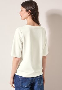 Top a maniche corte color crema chiaro con vestibilità rilassata, caratterizzato da un collo rotondo e un logo leggermente in rilievo sul retro. Finitura in tessuto texturizzato.