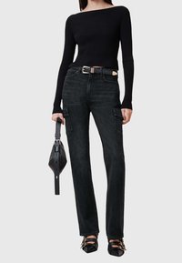 Haut noir à manches longues côtelé associé à un pantalon cargo noir taille haute avec poches latérales. Le mannequin tient un petit sac noir et porte des chaussures noires.