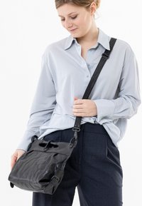 Schwarze Leder-Crossbody-Tasche mit strukturiertem Oberflächen, ausgestattet mit einem kurzen Tragegurt und Schlaufenakzenten. Das Model trägt ein hellblaues Hemd und dunkle Hosen.