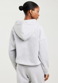 Kobieta o ciemnej karnacji i włosach upiętych w kok, ubrana w jasnoszary kaptur i dopasowane spodnie dresowe, stojąca bokiem z ręką swobodnie opuszczoną wzdłuż ciała.