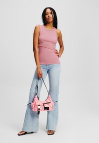 Lichtroze mouwloze top met asymmetrisch ontwerp en zwart logo, gecombineerd met lichtblauwe flared jeans en een handtas met een patroon in roze en wit.