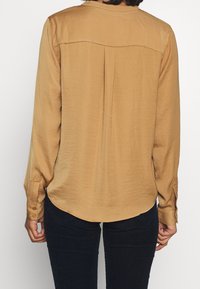 Long-sleeve, light brown blouse with a smooth, silky texture, featuring a yoke at the back and curved hem. Buttons at wrist cuffs.
