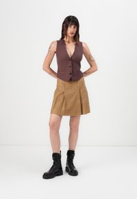 Gilet ajusté marron avec un décolleté plongeant, associé à une jupe plissée à carreaux jaunes et marron. Le modèle porte des bottines noires à lacets.