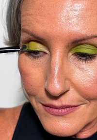 Dettaglio di una persona che applica ombretto verde brillante con un pennello. La pelle è liscia con un tono naturale e le labbra sono di un morbido mauve.