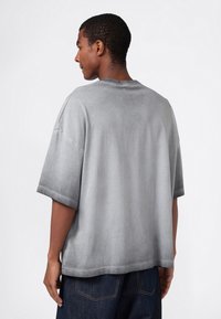 Junger Mann mit dem Rücken zur Kamera, der ein locker sitzendes graues T-Shirt mit kurzen Ärmeln und dunkle blaue Jeans trägt, vor einem schlichten weißen Hintergrund.