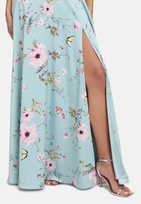 Abito maxi floreale di un blu chiaro con motivi rosa e verdi, caratterizzato da uno spacco alto fino alla coscia e abbinato a sandali con tacco alto e cinturini metallici.
