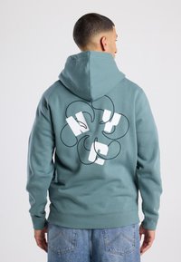 Trekker met capuchon in teal, voorzien van witte grafische tekst op de achterkant. Zachte textuur, geribbelde manchetten en een ontspannen pasvorm.