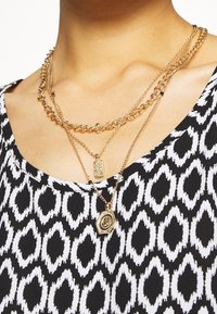 Zlaté vrstvené náhrdelníky s různými designy řetězů, včetně vzorovaného přívěsku a oválného šarmu, na černobíle vzorované látce.