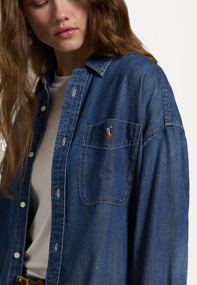Camicia di jeans blu scuro con colletto button-down, maniche lunghe e taschino sul petto con un piccolo logo ricamato. Texture liscia.