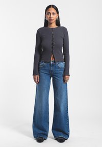 Top gris de manga larga con textura acanalada, con botones en el frente y diseño recortado, combinado con jeans de mezclilla azul de pierna ancha y zapatos negros.