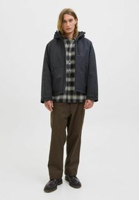 Chaqueta con capucha de color gris oscuro con cremallera en el delantero, dos bolsillos laterales, llevada sobre una camisa de cuadros de franela y combinada con pantalones sueltos marrones y zapatos negros.