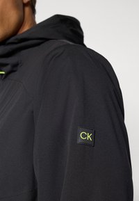 Svart jakke med hette, laget av et teksturert stoff. En firkantet CK-logo i neongrønn er brodert på venstre erm.