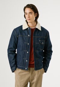 Chaqueta de mezclilla con cuello de borrego, color azul oscuro, botones de latón, bolsillo delantero, complementada con un suéter rojo debajo.