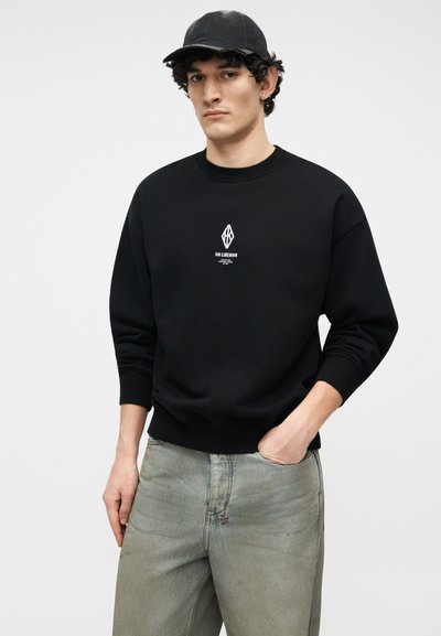 Νεαρός άνδρας που φοράει μαύρο καπέλο και μαύρο φούτερ με λογότυπο, ποζάροντας με το ένα χέρι στην τσέπη και ουδέτερη έκφραση.