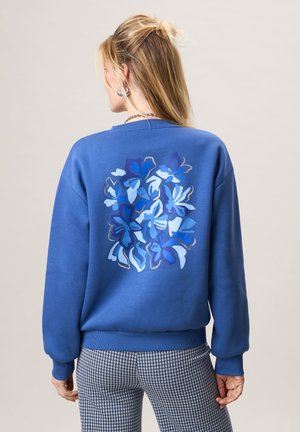 Vrouw draagt een blauw sweatshirt met een blauw bloemmotief op de rug en zwart-witte pied-de-poule broek, gezien van achteren.