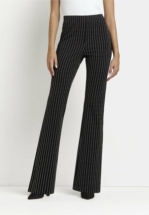 Žena v černých černobílých vysoce pasových kalhotách se zvonovým střihem, doplněných černými botami s špičatou špičkou, a bílém tílku bez rukávů.