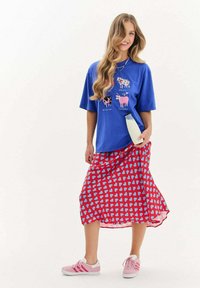 Modrá bavlnená tričko s grafikou kravy, skombinované s červenou sukňou s svetlomodrými srdiečkami. Modelka má na sebe ružové tenisky a drží fľašu mlieka.