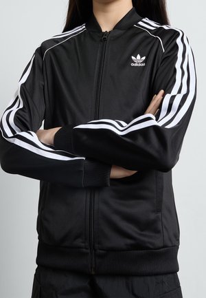 Personne les bras croisés portant une veste de survêtement Adidas noire avec des bandes blanches sur les manches et un logo sur la poitrine.