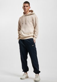 Felpa con cappuccio beige con parte superiore testurizzata e parte inferiore liscia, abbinata a pantaloni jogger blu scuro con tasche e dettagli a costine.