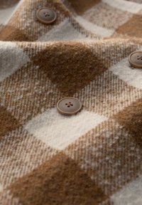 Braun- und cremefarbener Karostoff mit weicher Textur und einem braunen Knopf mit vier Löchern, der auf die Wolle oder eine Wollmischung hinweist.