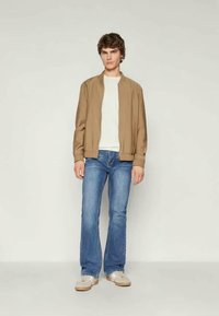 Veste bomber marron sur un t-shirt blanc, associée à un jean évasé bleu et des baskets claires. Tissu lisse et coupe décontractée.