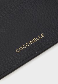 Czarny skórzany portfel na karty o teksturowanej powierzchni z złotym tłoczonym logo "COCCINELLE" w prawym dolnym rogu oraz widocznymi detalami szycia.