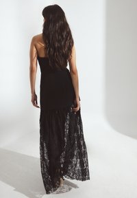 Moteris su ilgais tamsiais plaukais, vilkinti be rankovių juodą su nėriniu sijoną, eina tolyn prieš paprastą baltą foną.