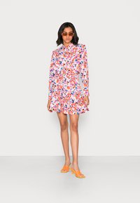 Vestido com padrão floral, mangas longas, barra com folhos e cintura ajustada. As cores incluem rosa, laranja, azul e verde. O modelo usa saltos laranja.