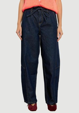 Frau trägt dunkelblaue locker sitzende Jeans mit Kordelzug in der Taille, rote Schuhe und einen hellen korallenfarbenen Pullover.