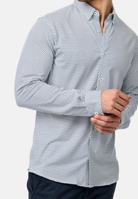 Košile na knoflíky s bílým základem a vzorem tmavě modrých geometrických trojúhelníků. Dlouhé rukávy, hladká texturu a standardní límec.