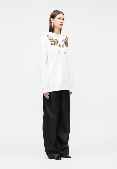 Γυναίκα φορώντας λευκό φούτερ με κέντημα φτερού τίγρη, μαύρο παντελόνι με φαρδιά γραμμή και μυτερά μαύρα παπούτσια, στέκεται μπροστά σε απλό λευκό φόντο.