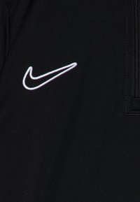 Czarna tkanina z haftowanym, świecącym białym i fioletowym logo Nike w kształcie łyżwy, umieszczonym obok czarnego zamka błyskawicznego po prawej stronie.
