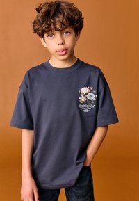 Sötétszürke, rövid ujjú póló a bal mellrészen koponya és virág motívummal, pamutból, sima textúrával.